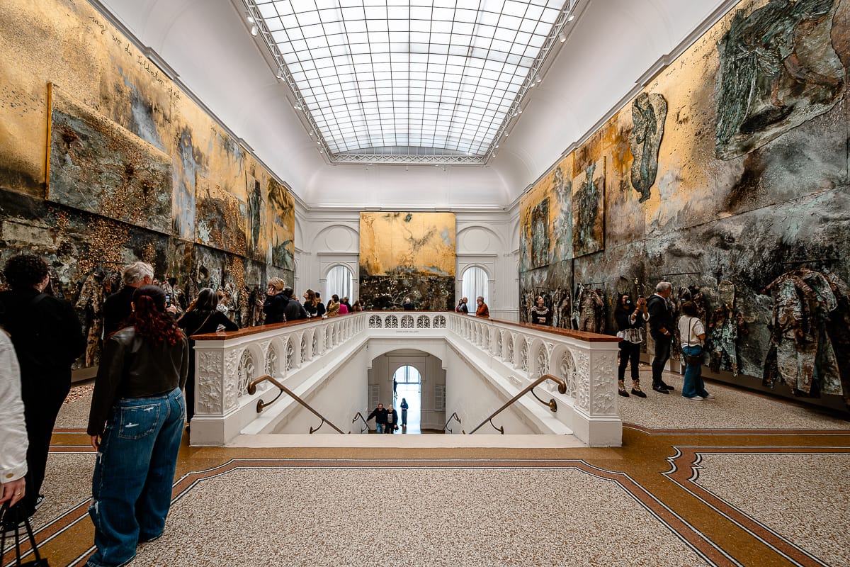 Eine geräumige Kunstgalerie mit hohen Decken, Oberlichtern und großen abstrakten goldfarbenen Gemälden. Besucher stehen und gehen umher und betrachten die Kunstwerke. Eine verzierte weiße Treppe führt hinunter zu einem hellen Eingang am anderen Ende.