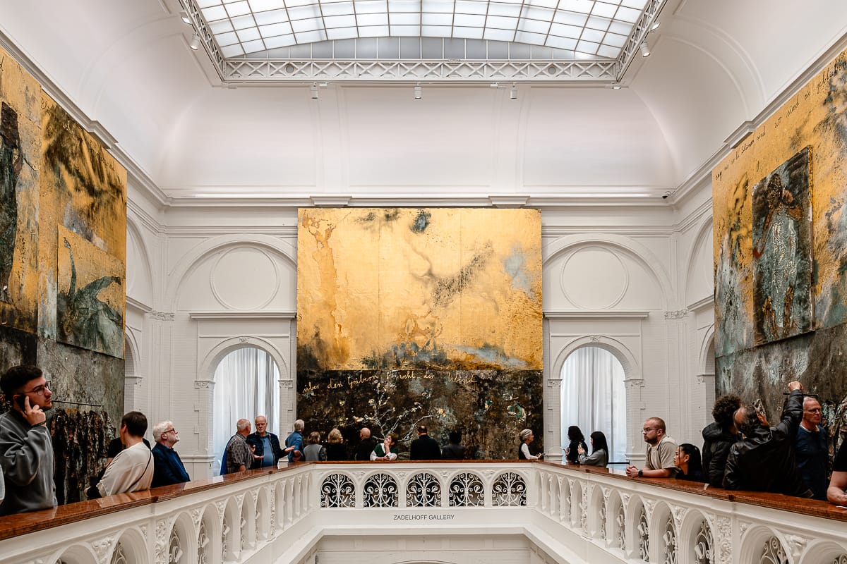 Eine Gruppe von Menschen betrachtet große abstrakte Gemälde in Gold und dunklen Tönen von Anselm Kiefer in einer hellen Kunstgalerie mit Glasdecke und verziertem weißen Geländer. Das zentrale Kunstwerk dominiert die Wand, während Besucher beobachten, sich unterhalten und Fotos machen. Der Raum wirkt offen und elegant.