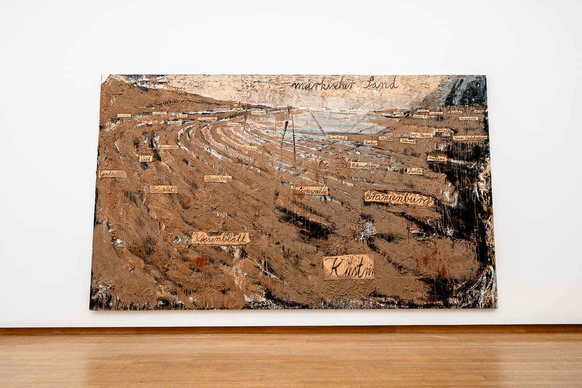 Ein großes Gemälde von Anselm Kiefer an einer weißen Galeriewand zeigt braune, erdige Texturen mit abstrakten, geschwungenen Linien und rauen Stellen. Rechteckige Beschriftungen mit deutschen Wörtern sind über die Landschaft verteilt und erinnern an eine topografische Karte. Der Boden ist aus Holz.