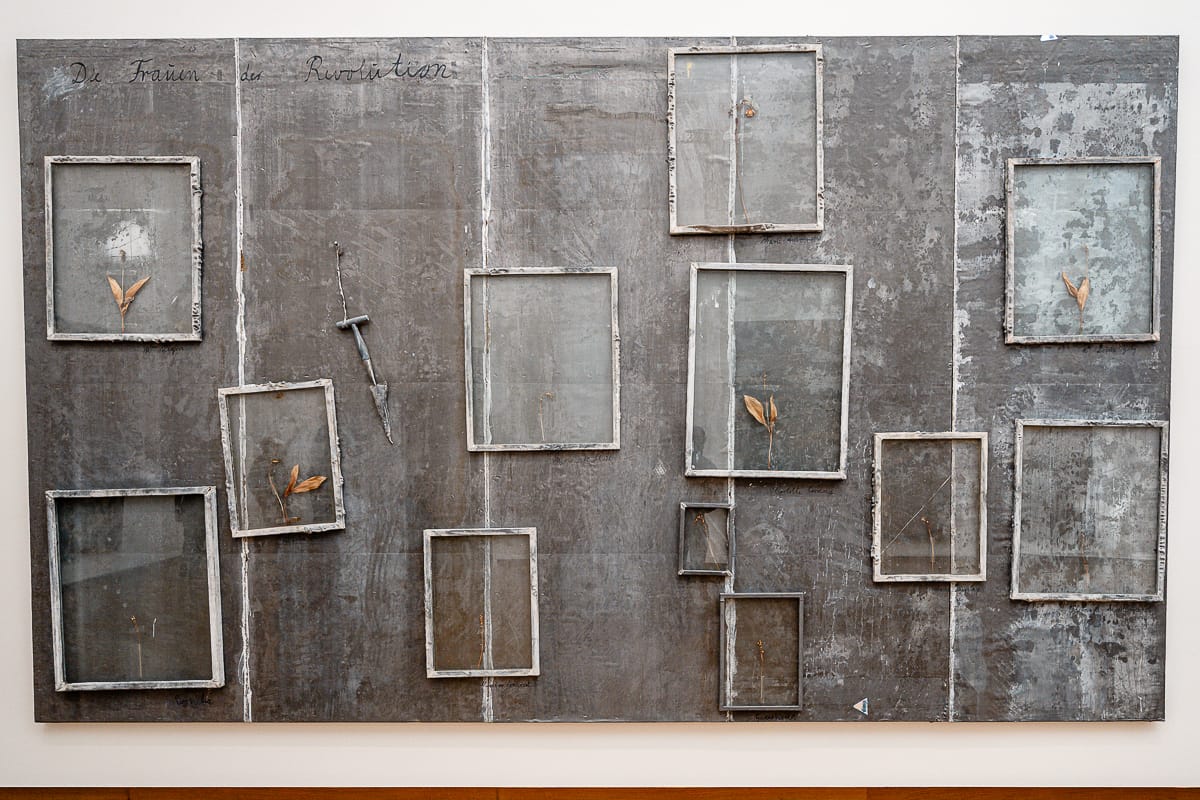 Ein großes, grau getöntes Kunstwerk von Anselm Kiefer zeigt leere, unregelmäßig angeordnete Bilderrahmen unterschiedlicher Größe, einige davon schräg. Mehrere Rahmen enthalten getrocknete Blätter. Der strukturierte Hintergrund erinnert an verwitterten Beton, und oben links ist eine schwache Handschrift zu sehen.