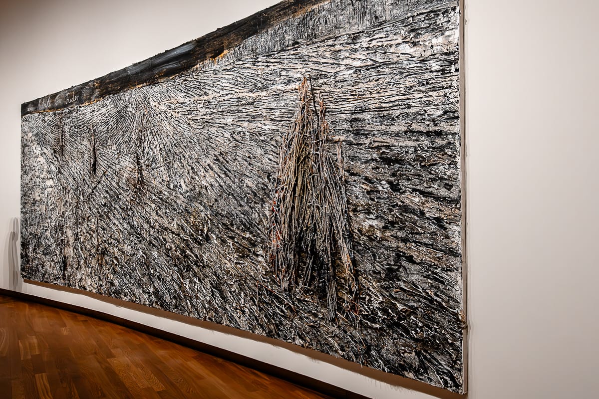 Ein großes, strukturiertes abstraktes Gemälde  von Anselm Kiefer dominiert eine Galeriewand mit dunklen Erdtönen und dicken, taktilen Schichten. Die Komposition zeugt von rauen, strahlenförmigen Linien und zentralen Graten und erinnert an eine zerklüftete Landschaft. Der Boden ist aus Holz, die Wand weiß.
