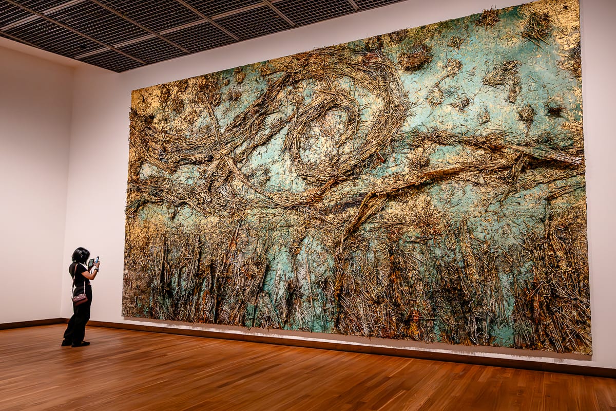 Eine Person steht in einer Galerie und fotografiert ein großes strukturiertes Kunstwerk - De sterrennacht (The Starry Night) von Anselm Kiefer -  mit wirbelnden Mustern in Gold und Braun auf blaugrünem Hintergrund. Das Mixed-Media-Werk ist an einer weißen Wand montiert und reicht vom Boden bis zur Decke, sodass der Betrachter winzig wirkt.