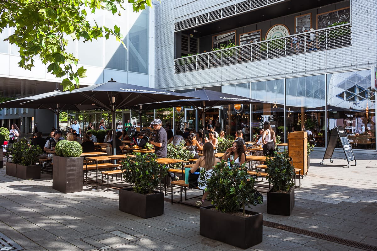 Café-Szene im Freien mit Holztischen und Bänken unter großen schwarzen Schirmen. Die Menschen sitzen, essen und unterhalten sich im Frittenwerk München. Grüne Topfpflanzen schmücken den Bereich, mit einem modernen Glas- und Backsteingebäude im Hintergrund an einem hellen, sonnigen Tag.