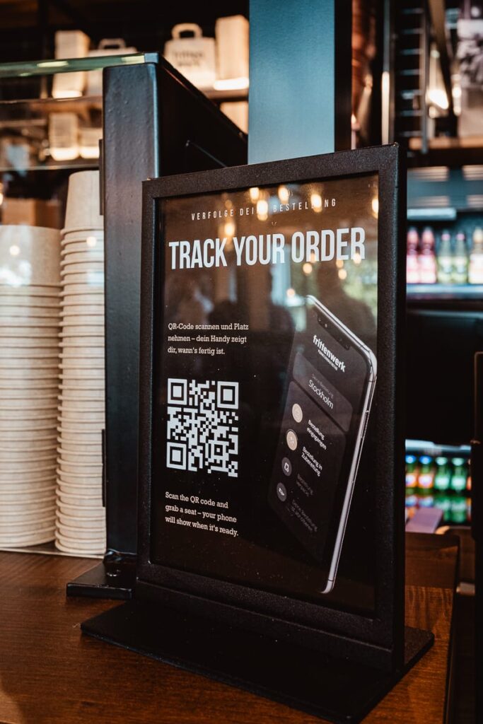 Ein Schild an der Theke im Frittenwerk München zeigt "Track Your Order" über einem QR-Code und einem Telefonbild. Die Kunden werden eingeladen, den Code zu scannen, um den Status ihrer Bestellung zu überprüfen.