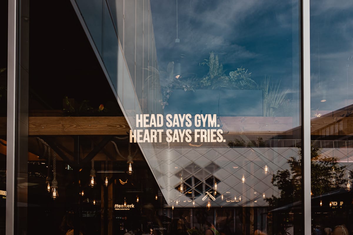 Ein Glasfenster im Frittenwerk München zeigt den Text "HEAD SAYS GYM. HEART SAYS FRIES." Spiegelungen von Bäumen, Gebäuden und Innenbeleuchtung vermischen sich mit Ansichten von Pflanzen und hängenden Glühbirnen im Inneren der Einrichtung.
