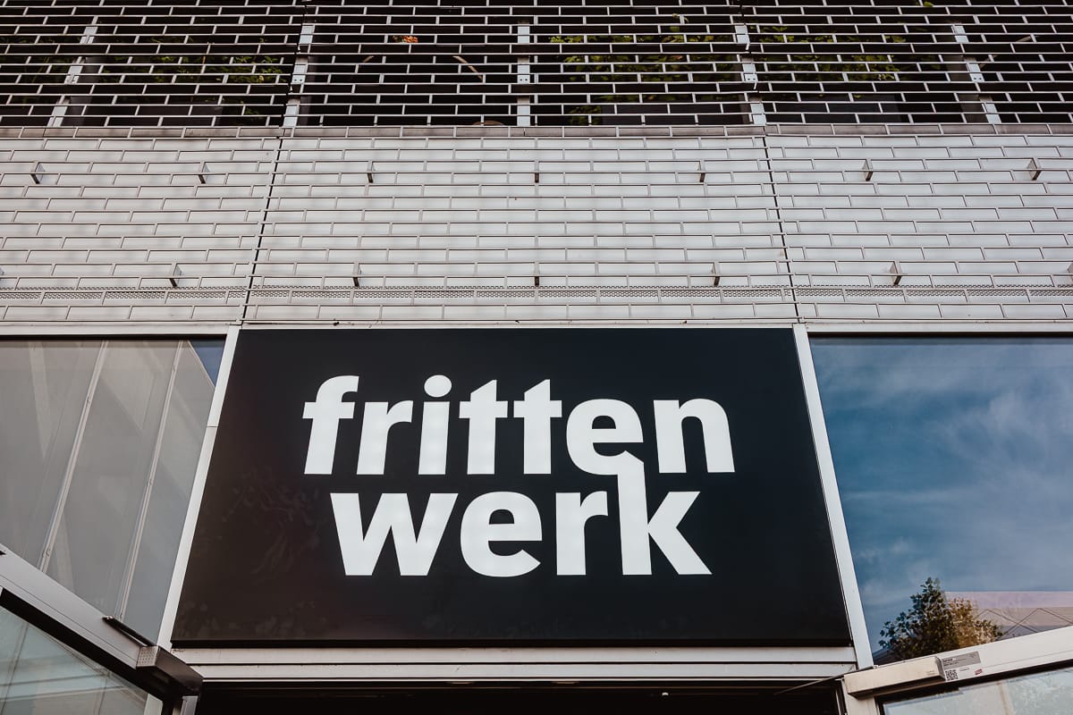 Ein schwarzes Schild mit großen, fetten weißen Buchstaben, auf dem "frittenwerk" steht, ist an einer weißen Backsteinwand am Frittenwerk München angebracht. Oben zeigen Metallstangen kleine grüne Pflanzen, während das Glas darunter den Himmel und einen Teil eines Gebäudes reflektiert.