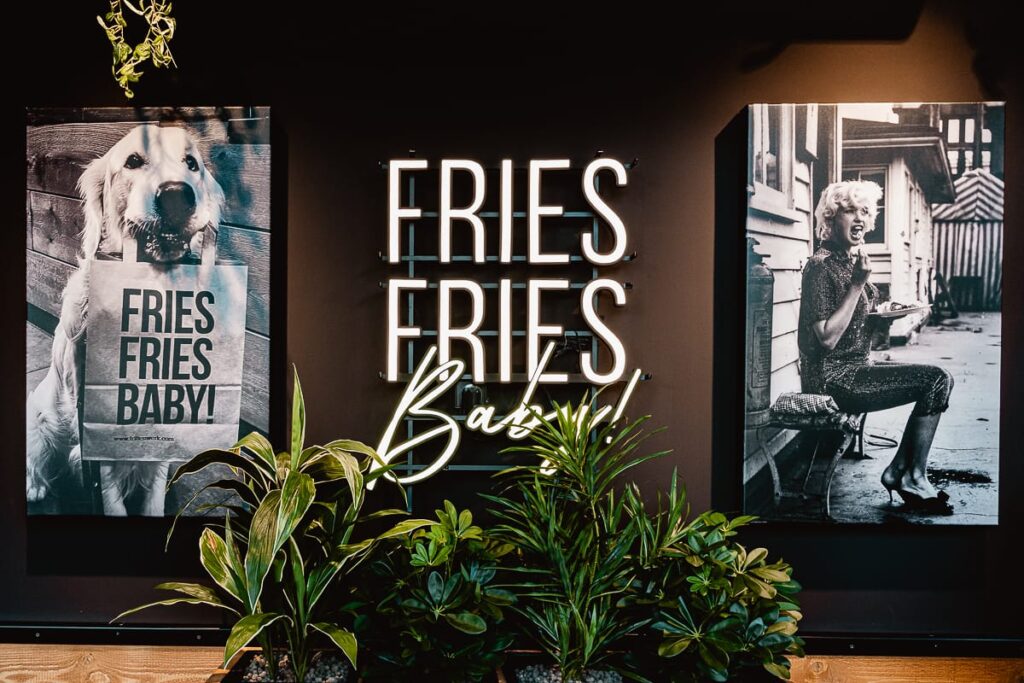 Eine Leuchtreklame mit der Aufschrift "FRIES FRIES Baby" im Frittenwerk München, ist an einer dunklen Wand angebracht. Links hält ein Hund eine "FRIES FRIES BABY!"-Tüte. Rechts zeigt ein Schwarz-Weiß-Foto eine Frau, die über grünen Blattpflanzen Pommes isst.