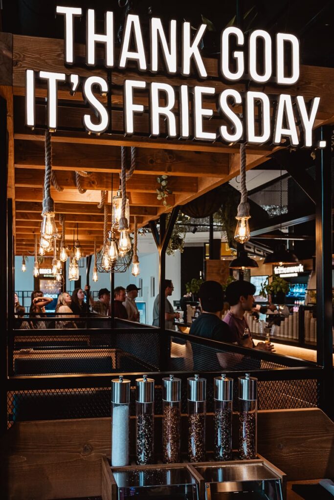 Im Frittenwerk München leuchtet ein Schild mit der Aufschrift "THANK GOD IT'S FRIESDAY". Hängende Glühbirnen und Holzbalken schmücken die Decke, während sich die Menschen in einer warmen, lebendigen Atmosphäre tummeln. Unter der Leuchtschrift sieht man Gewürzspender, die Teil der Self Service Station sind.
