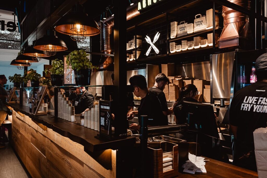 Im Frittenwerk München, einem modernen, schummrig beleuchteten Restaurant mit Holz- und Metalldekor, arbeiten mehrere Personen hinter der Theke. Pflanzen und gestapelte Papiertüten sind zu sehen, während das Personal Speisen und Getränke zubereitet und ein Kunde in einem T-Shirt mit Slogan an der Theke wartet.