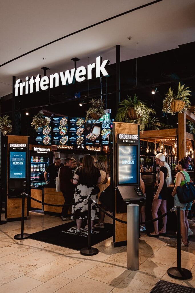 Eine Gruppe von Menschen steht in der Schlange im Frittenwerk München, einem Restaurant mit einem leuchtenden Schild, digitalen Menütafeln und von der Decke hängenden Pflanzen. Zwei Touchscreen-Bestellkioske begrüßen die Gäste am Eingang, während Holzakzente und bunte Beleuchtung den Raum aufwerten.