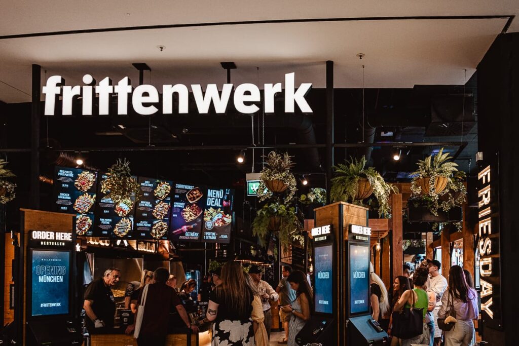 Ein belebtes Restaurant namens "Frittenwerk München" mit einer großen Leuchtreklame. Die Leute stehen Schlange, um Essen zu bestellen. Hängende Pflanzen schmücken die Decke, und Bildschirme zeigen die Speisekarte an. Die Atmosphäre ist lebendig und modern.