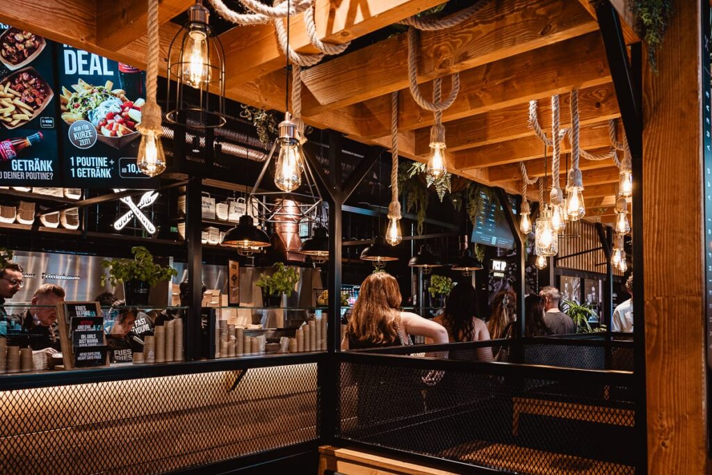 Ein modernes Restaurant mit Holzbalken, freiliegenden Glühbirnen und hängenden Pflanzen. Mehrere Personen sitzen an Tischen. Der Thekenbereich im Frittenwerk München zeigt digitale Menütafeln mit Speiseangeboten und Angeboten. Die warme, einladende Beleuchtung sorgt für die richtige Stimmung.