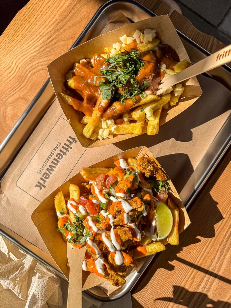 Zwei Tabletts mit gefüllten Pommes frites von Frittenwerk München liegen auf einem Metalltablett. Auf dem oberen Tablett befinden sich Pommes frites mit brauner Soße, Käsebruch und Kräutern, auf dem unteren gebratenes Hähnchen, Gemüse, Soßen und eine Limettenspalte. In jedes Tablett sind Gabeln eingesteckt.
