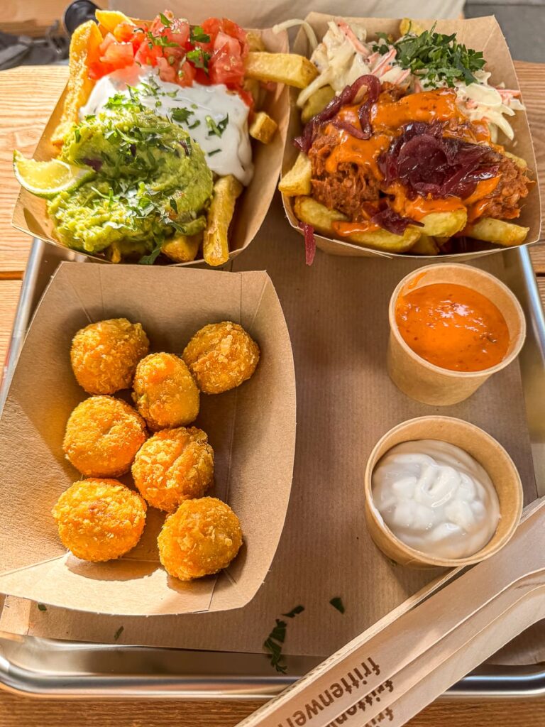 Auf einem Metalltablett im Frittenwerk München liegen gefüllte Pommes frites mit Guacamole, Sauerrahm, Tomaten, Pulled Meat, Krautsalat und Zwiebeln, knusprig frittierte Bällchen in einem Papierschiffchen und zwei Dip-Saucen auf einem Holztisch mit Holzbesteck.