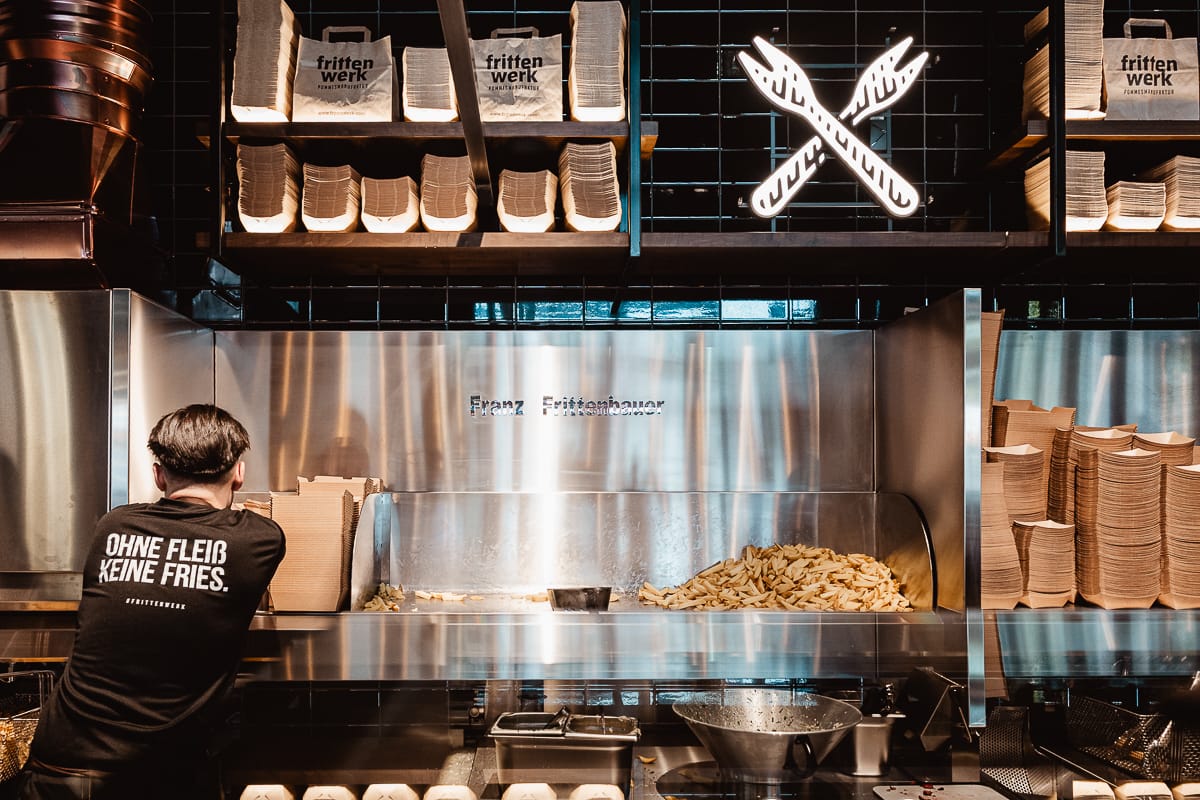 Ein Arbeiter im schwarzen Hemd steht im Frittenwerk München vor einer mit Pommes gefüllten Edelstahltheke, darüber stapeln sich Pappschalen und Tüten mit der Aufschrift "frittenwerk". Ein Neonschild mit gekreuzten Gabeln und Messern leuchtet an der dunkel gekachelten Wand.