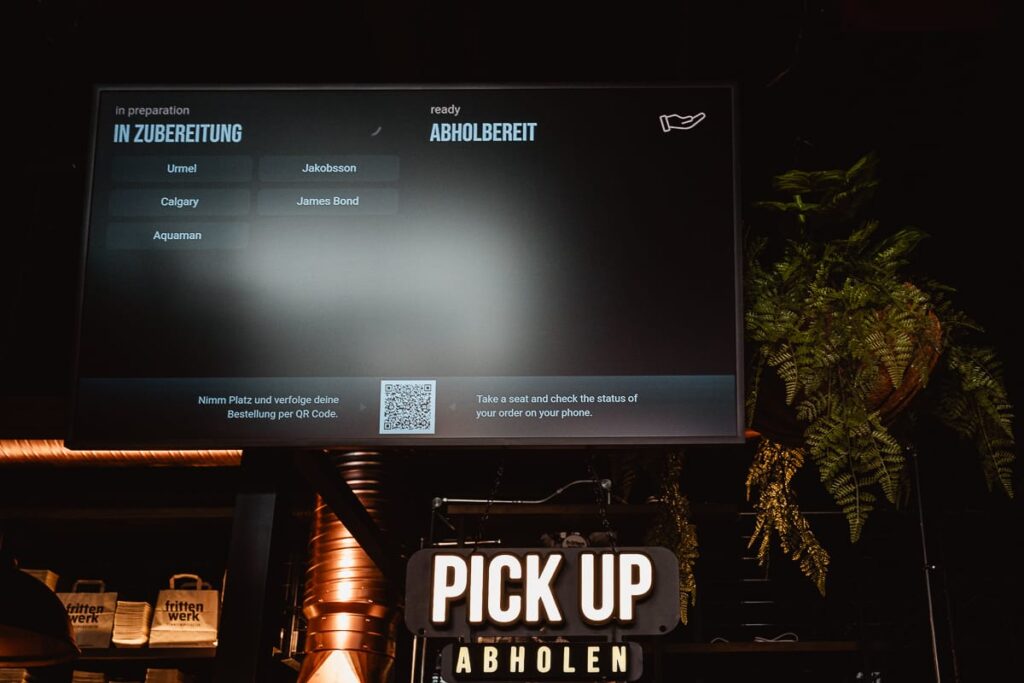 Ein Bildschirm im Frittenwerk München zeigt den Bestellstatus auf Deutsch und Englisch an, mit Namen wie Jakobsson, James Bond und Aquaman. Darunter leuchtet ein helles "PICK UP ABHOLEN"-Schild, während in der Nähe eine grüne Blattpflanze hängt und im Hintergrund schummrige Regale stehen.