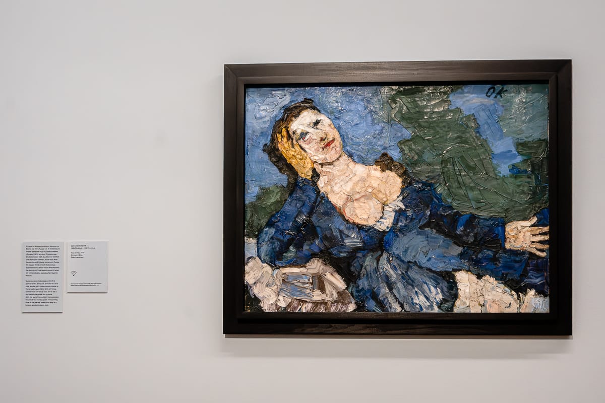 Ein strukturiertes Gemälde einer liegenden Frau - die Alma Mahler nachempfunde Puppe - in einem blauen Kleid mit tiefem Ausschnitt, das mit ausdrucksstarken, dicken Pinselstrichen wiedergegeben ist. Das Bild ist schwarz gerahmt und hängt an einer weißen Wand neben zwei kleinen Informationsschildern. Der Hintergrund ist überwiegend blau und grün.