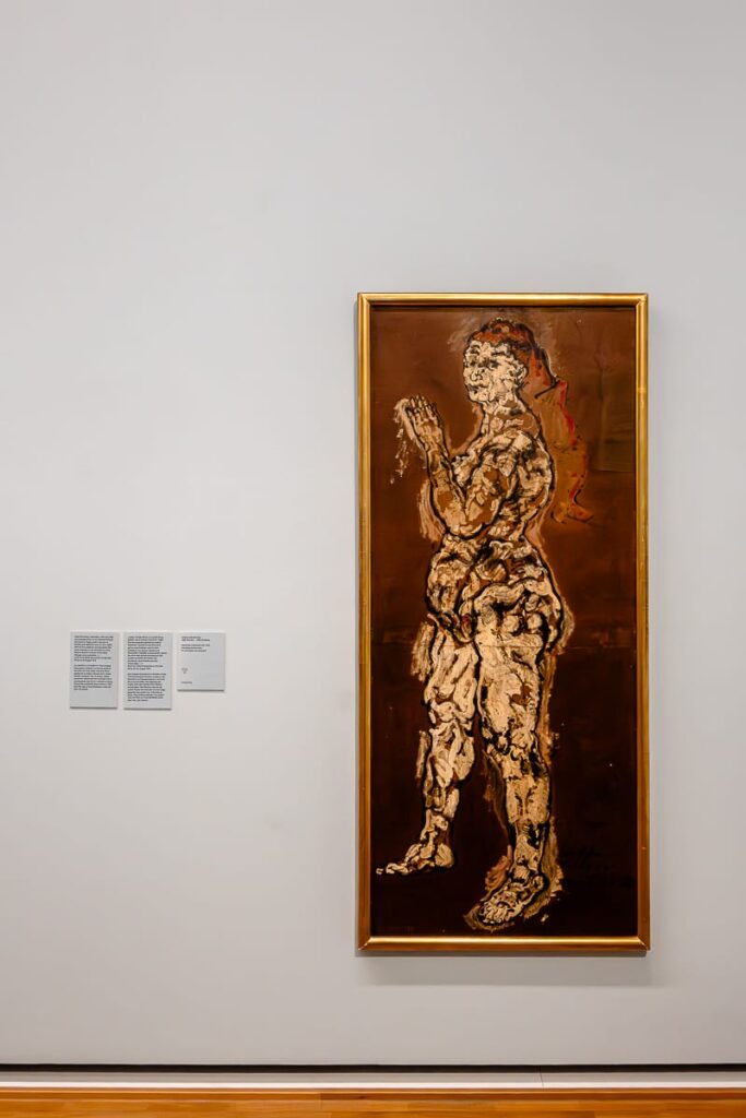 Ein gerahmtes Gemälde einer stehenden menschlichen Figur, eine stilisierte Alma Mahler, mit groben, strukturierten Pinselstrichen in Creme-, Beige- und Brauntönen auf dunkelbraunem Hintergrund hängt an einer hellgrauen Galeriewand, neben zwei Wandschildern mit Text. Die Figur erscheint abstrakt und blickt nach oben.