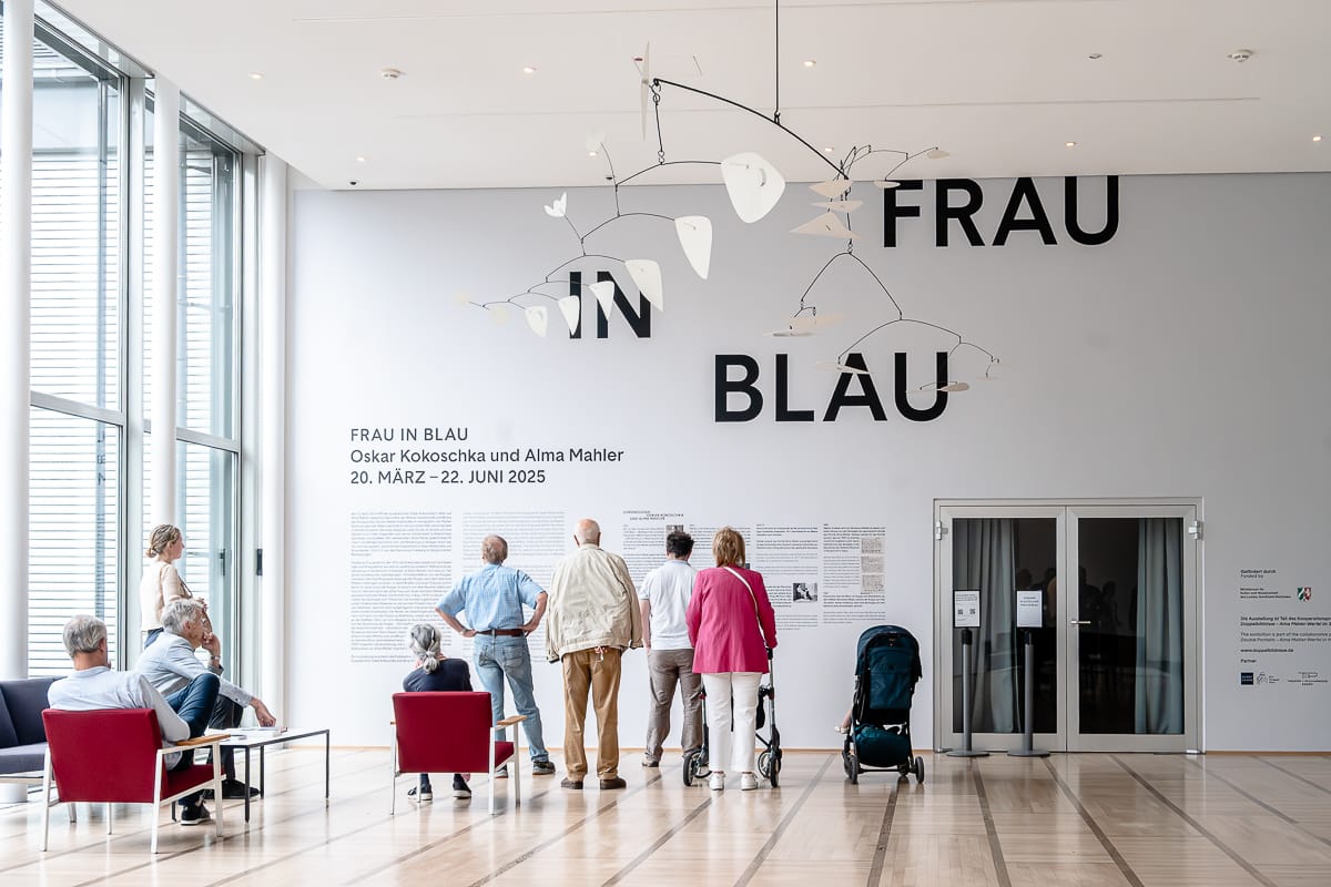 Eine Gruppe von Menschen steht und sitzt in einer modernen Museumshalle mit großen Fenstern. An der Wand steht in fetter Schrift "FRAU IN BLAU". Die Ausstellungsinformation bezieht sich auf Alma Mahler. Ein großes weißes Mobile hängt von der Decke, und ein Kinderwagen steht in der Nähe.