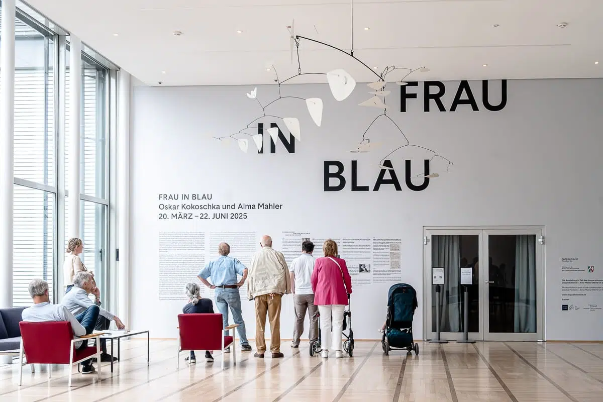 Eine Gruppe von Menschen steht und sitzt in einer modernen Museumshalle mit großen Fenstern. An der Wand steht in fetter Schrift "FRAU IN BLAU". Die Ausstellungsinformation bezieht sich auf Alma Mahler. Ein großes weißes Mobile hängt von der Decke, und ein Kinderwagen steht in der Nähe.