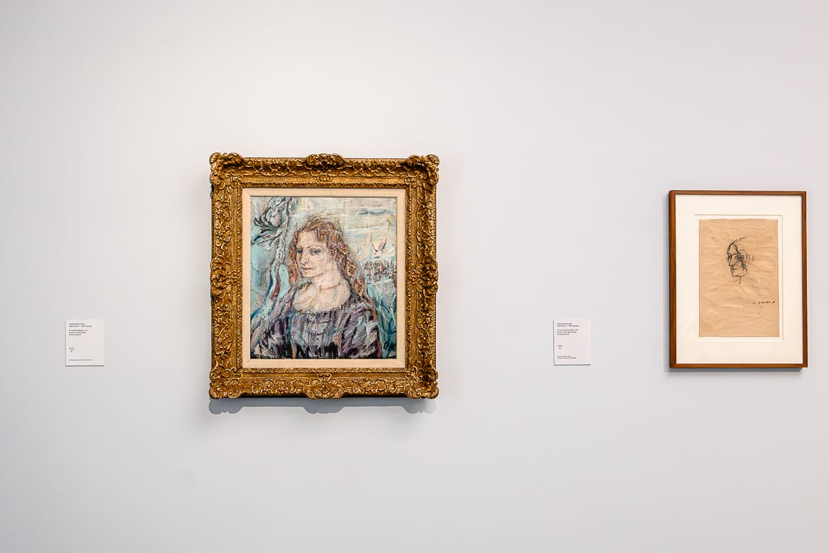 An einer weißen Wand hängt ein verziertes, goldgerahmtes Gemälde einer Frau mit gewelltem Haar und düsterem Gesichtsausdruck - Alma Mahler, gezeichnet von Oskar Kokoschka. Daneben ist eine einfache, braun gerahmte Bleistiftskizze eines Kopfes auf hellbraunem Papier zu sehen. Neben jedem Kunstwerk sind kleine weiße Plakate angebracht.