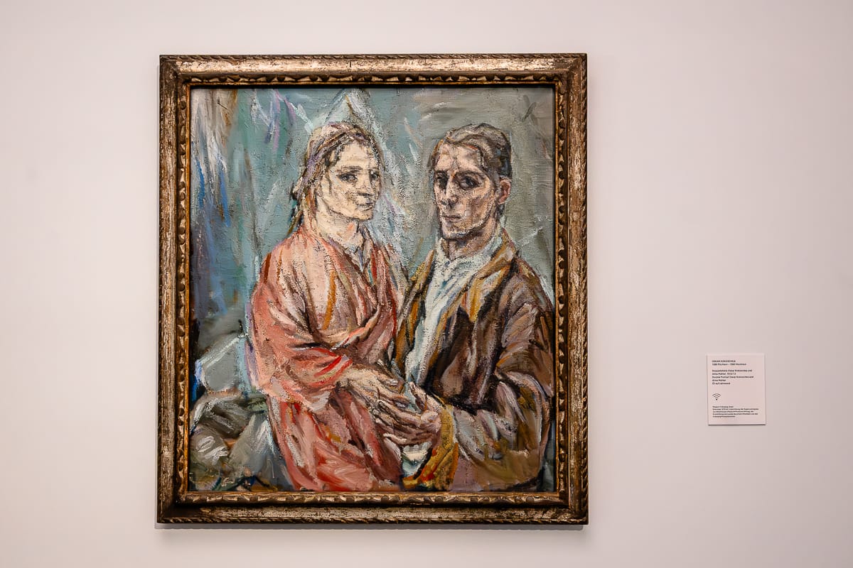 An einer weißen Wand hängt ein gerahmtes Gemälde, auf dem ein Mann (Oskar Kokoschka) und eine Frau (Alma Mahler) mit ernster Miene zu sehen sind. Die Frau trägt ein rosafarbenes Gewand, der Mann eine braune Jacke. Ihre Hände berühren sich sanft. Der Stil ist ausdrucksstark mit kräftigen Pinselstrichen und gedämpften Farben.