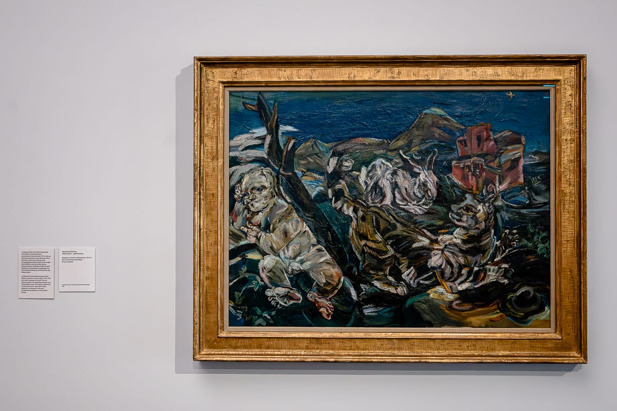 An einer weißen Wand hängt ein gerahmtes abstraktes Gemälde, das verzerrte tierähnliche Figuren in dynamischen Posen vor einer dunklen Berglandschaft zeigt. Links daneben beschreiben zwei kleine weiße Museumsschilder das Kunstwerk und den Künstler.