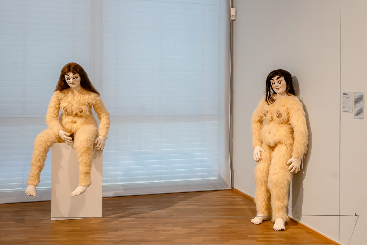Zwei menschenähnliche Skulpturen (Puppen nach der Alma-Puppe von Kokoschka) mit hellgelbem Fell stehen in einer Galerie mit weißen Wänden und Holzböden; die eine sitzt auf einem weißen Sockel, die andere lehnt an der Wand. Beide haben weiße Hände, Füße und Gesichter mit braunem Haar und neutralem Gesichtsausdruck.