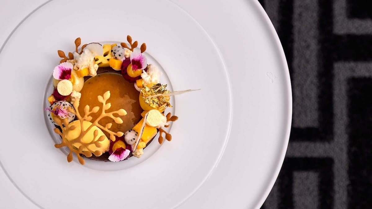 Ein Gourmet-Dessert liegt auf einem großen weißen Teller. Das runde Gebäck ist mit glänzender Karamellglasur, Blattgold, kleinen Baisers, essbaren Blumen und kunstvoll arrangierten Schokoladendekorationen überzogen. Der Hintergrund hat eine schwarze, geometrisch gemusterte Oberfläche.