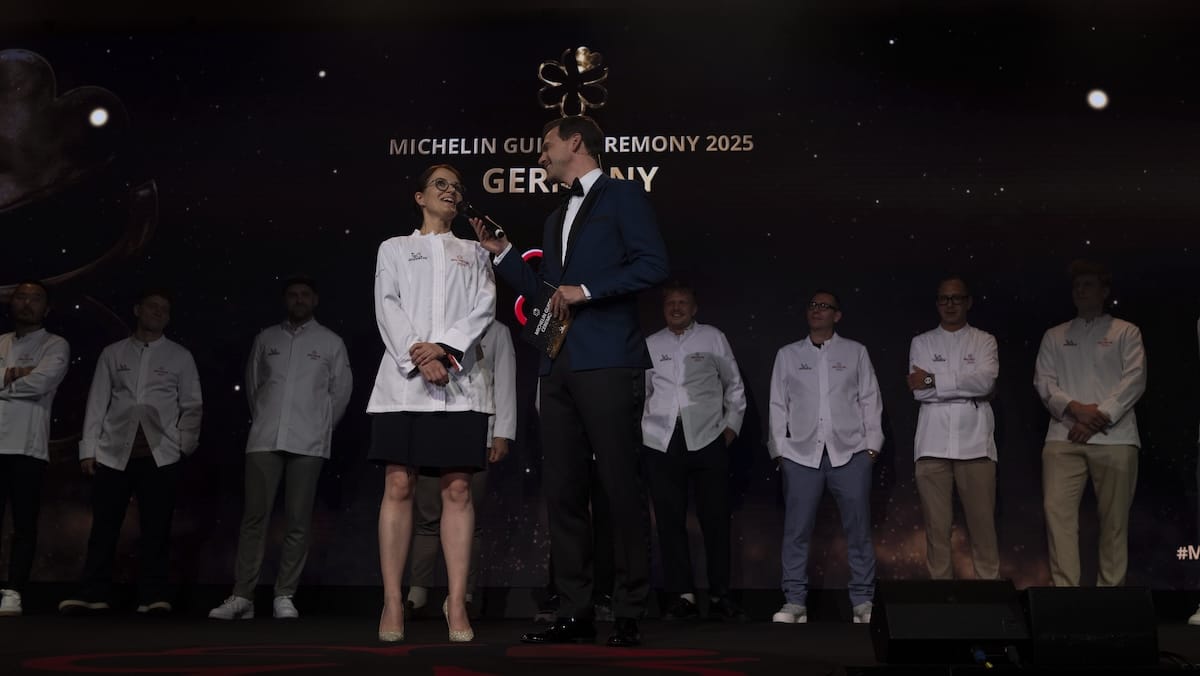 Eine Gruppe von Köchen in weißen Jacken steht auf einer dunklen Bühne während der Verleihung des Michelin Guides 2025 in Deutschland. Eine Frau und ein Mann, die beide ein Mikrofon in der Hand halten, werden in der Mitte angestrahlt, während sechs weitere Köche im Hintergrund stehen.