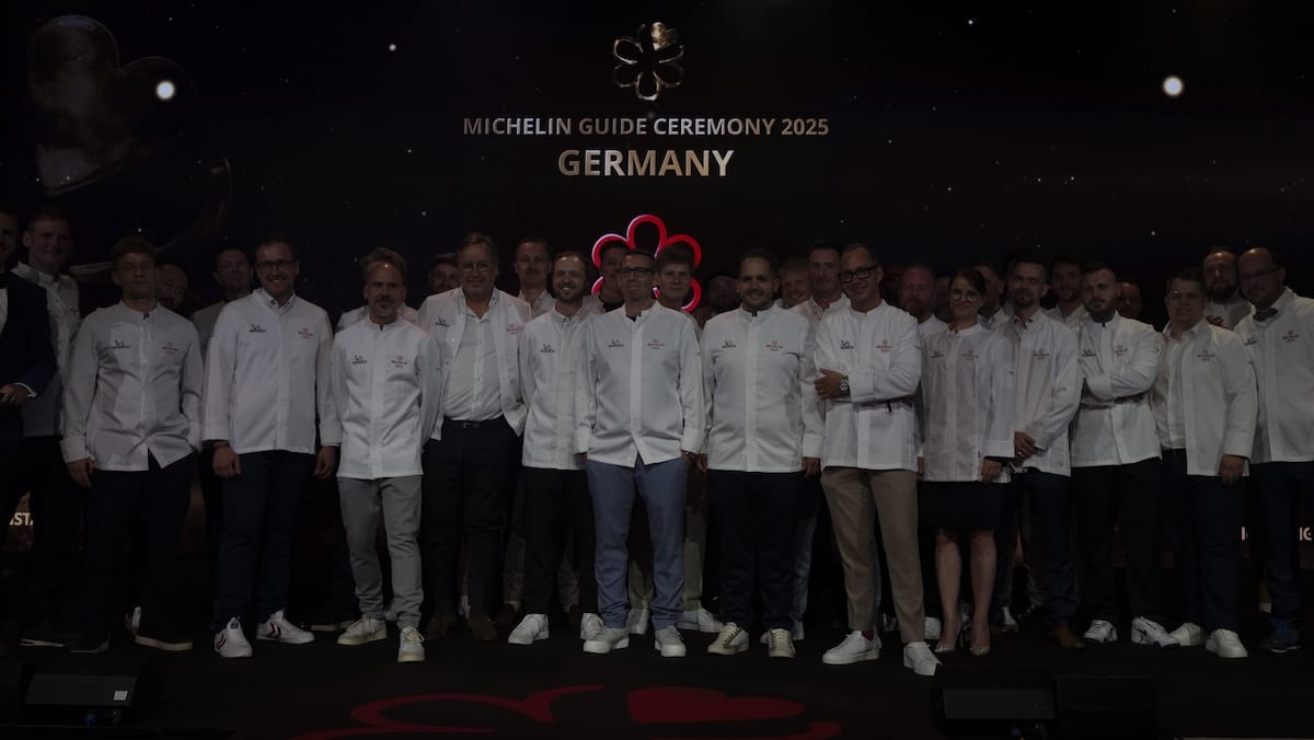 Eine Gruppe von Köchen in weißen Jacken steht bei der Verleihung des Michelin Guide 2025 Deutschland gemeinsam auf der Bühne und lächelt vor einem dunklen Hintergrund, über dem das Michelin-Logo und der Titel der Veranstaltung leuchten.