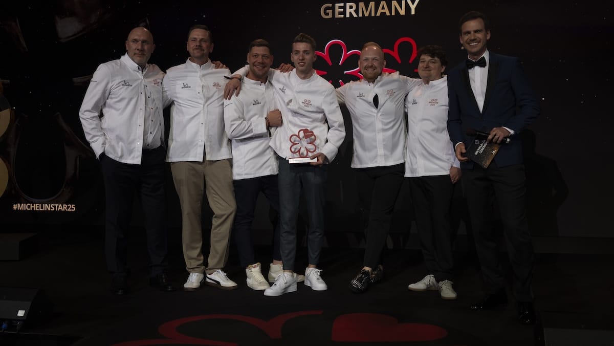 Sieben Personen stehen auf einer schwach beleuchteten Bühne. Sechs tragen weiße Kochjacken und Freizeithosen, lächeln und posieren eng beieinander; einer hält eine Auszeichnung des Guide Michelin 2025 in der Hand. Die siebte Person in einem dunklen Anzug hält ein Mikrofon. Über ihnen erscheinen "GERMANY" und ein Michelin-Stern-Symbol.
