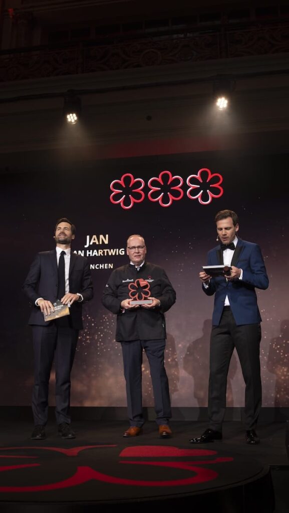 Drei Männer in formeller Kleidung stehen auf einer Bühne unter roten Michelin-Sternen. Der Mann in der Mitte hält eine große rote Auszeichnung des Michelin Guide 2025 in der Hand. Hinter ihnen zeigt ein dunkler Bildschirm "JAN" und "MÜNCHEN" an, die von zwei Scheinwerfern von oben angestrahlt werden.