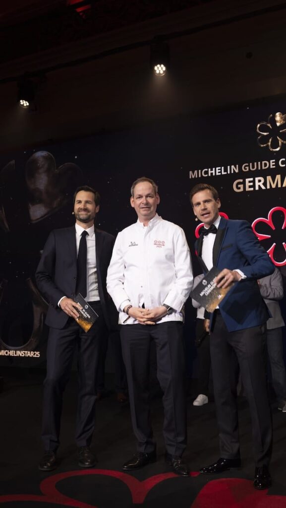 Drei Männer posieren auf der Bühne bei der Verleihung des Michelin Guides 2025 in Deutschland. Der Mann in der Mitte trägt eine weiße Kochjacke mit Michelin-Sternen, während die beiden Männer, die ihn flankieren, dunkle Anzüge tragen und Veranstaltungskarten halten. Im Hintergrund sind ein Schild des Michelin-Führers Deutschland und Sternsymbole zu sehen.