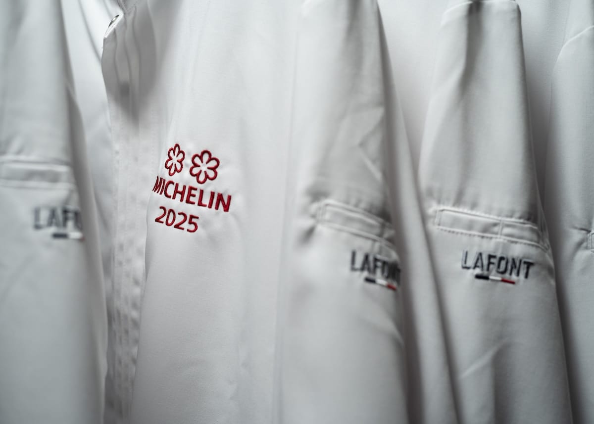 Nahaufnahme von weißen, hängenden Kochjacken, die auf der Tasche mit "LAFONT" bestickt sind. Auf einer Jacke prangt der Schriftzug "MICHELIN 2025" mit zwei Michelin-Sternen, der die Aufnahme in den Michelin Guide 2025 kennzeichnet.