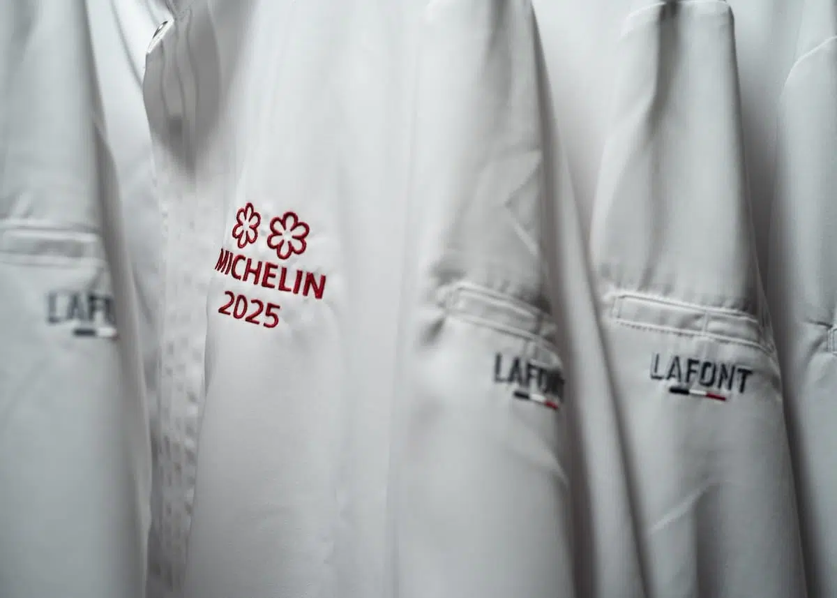 Nahaufnahme von weißen, hängenden Kochjacken, die auf der Tasche mit "LAFONT" bestickt sind. Auf einer Jacke prangt der Schriftzug "MICHELIN 2025" mit zwei Michelin-Sternen, der die Aufnahme in den Michelin Guide 2025 kennzeichnet.