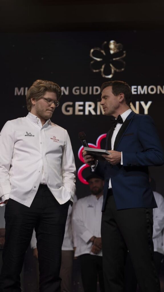 Ein Koch in einer weißen Jacke steht auf der Bühne neben einem Mann in einem blauen Anzug, der ein Mikrofon und einen Notizblock hält. Im Hintergrund steht "MICHELIN GUIDE CEREMONY GERMANY" mit Michelin-Sternen -Verleihung zum Michelin Guide 2025. Beide Männer scheinen konzentriert zu sein, möglicherweise während eines Interviews oder einer Preisverleihung.