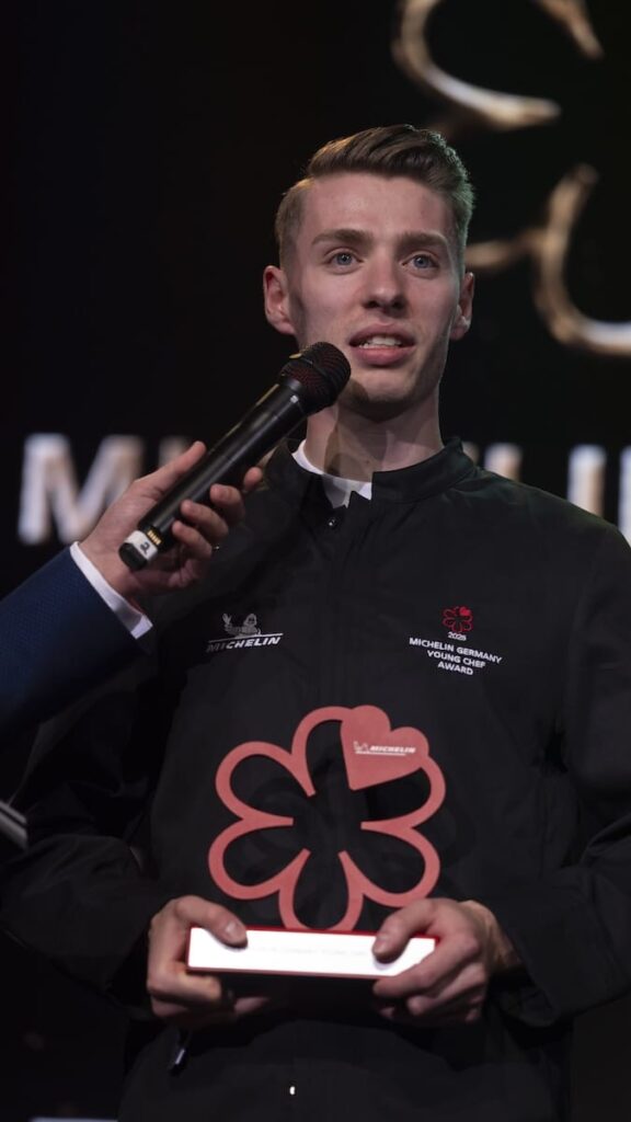 Ein junger Koch in einer schwarzen Jacke des Michelin Germany Awards hält eine rote Michelin-Stern-Trophäe. Er spricht in ein Mikrofon und steht vor einem dunklen Hintergrund mit verschwommenem Text, dem großen Michelin-Stern-Symbol und dem Michelin Guide 2025, auf den er stolz zeigt.