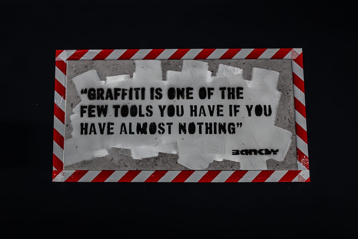 Auf einer rechteckigen Platte ist in schwarzer Schablone ein Banksy-Zitat eingraviert: "Graffiti ist eines der wenigen Werkzeuge, die man hat, wenn man fast nichts hat." Es ist teilweise mit weißer Farbe übermalt, mit "BANKSY" signiert und mit rot-weißen Warnstreifen umrandet.