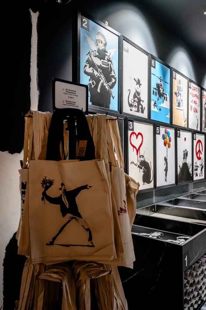 Vor einer Wand mit gerahmten Banksy-Drucken - Graffiti-Polizei, Ratten, das Ballonmädchen und Friedenssymbole - in einem hell erleuchteten, modernen Laden hängen Banksy-Taschen mit dem ikonischen Blumenwerfer.