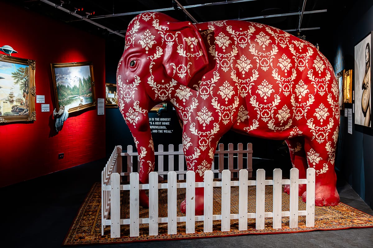 Eine lebensgroße rote Elefantenskulptur mit weißen Damastmustern steht hinter einem kleinen weißen Lattenzaun in einer Kunstgalerie mit einer Banksy-Ausstellung. Sie steht auf einem gemusterten Teppich unter dramatischer Beleuchtung, eingerahmt von dunkelroten Wänden.