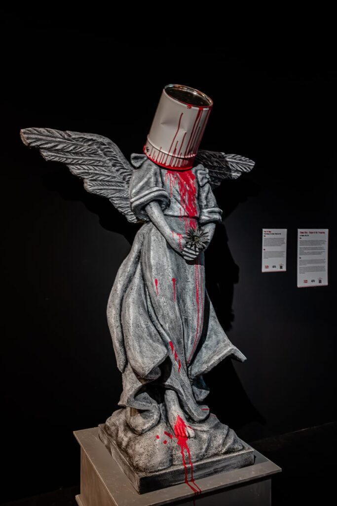 Eine graue Engelsstatue mit gefalteten Händen steht vor einem schwarzen Hintergrund. Ihr Kopf ist durch eine umgestürzte Farbdose ersetzt, von der rote Farbe heruntertropft und sich darunter sammelt. In der Nähe sind zwei Wandplakate zu sehen.