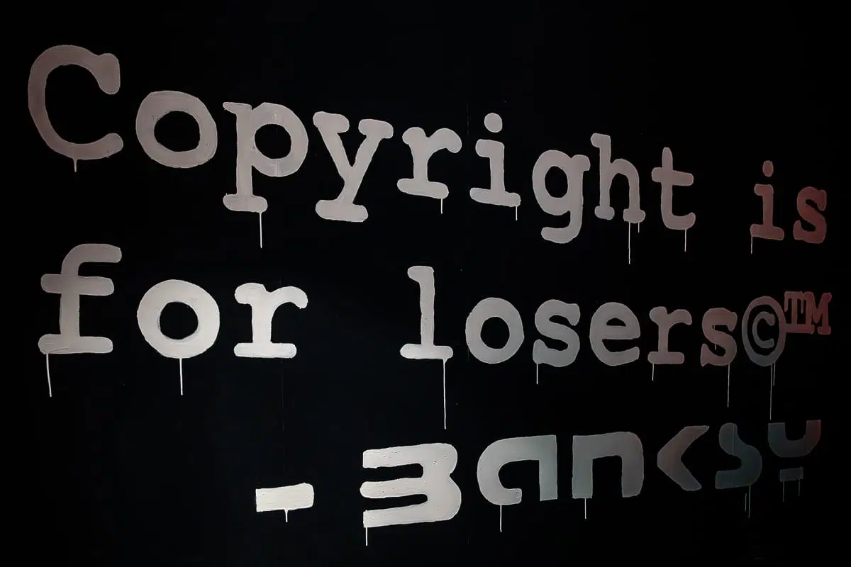 Ein großer, weißer Graffiti-Text auf einer schwarzen Wand lautet: "Copyright is for losers©™", wobei das Wort "Banksy" unten teilweise verdeckt ist. Die Farbe tropft leicht und erinnert an den rebellischen Street-Art-Stil von Banksy.