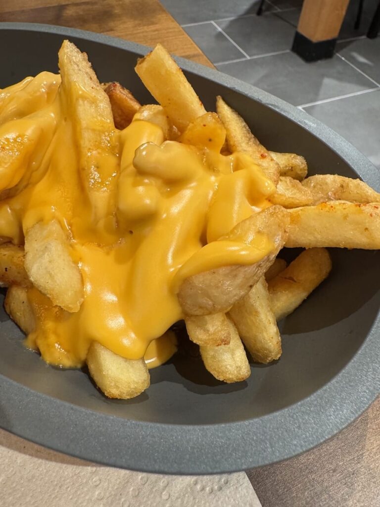 Ein schwarzer ovaler Teller von Frittenwerk Bielefeld ist mit dick geschnittenen, goldgelben Pommes frites gefüllt, die mit geschmolzener gelber Käsesauce überzogen sind. Die knusprigen Pommes frites sind großzügig bedeckt, und der Teller steht auf einem Holztisch mit grauem Kachelboden im Hintergrund.