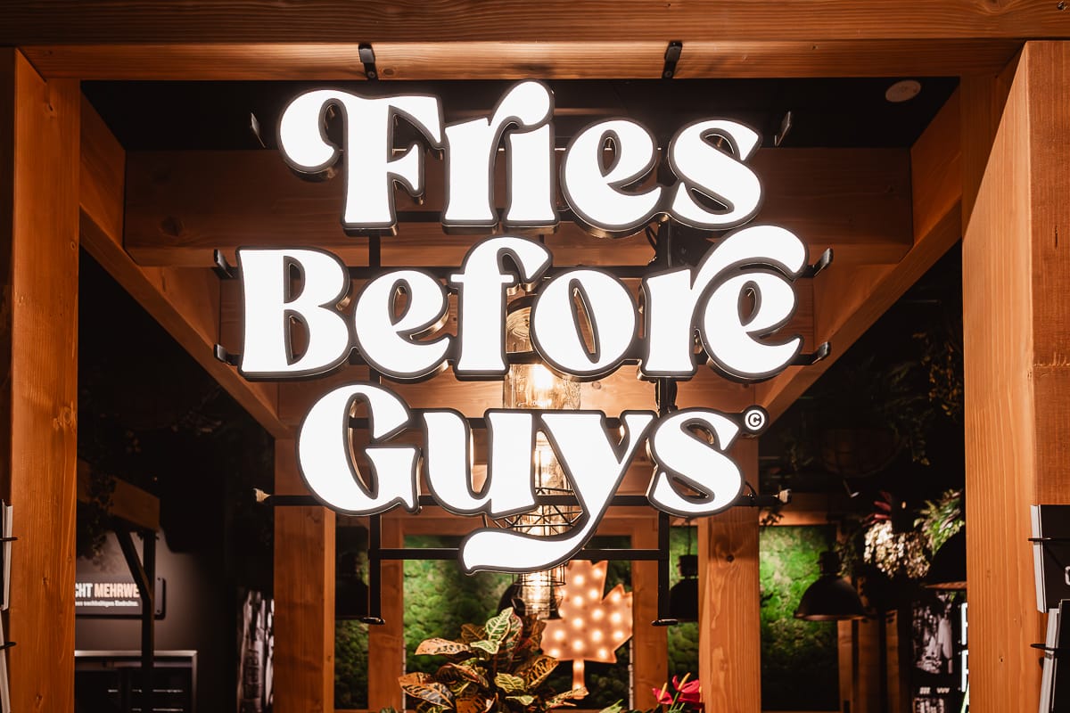 Ein großes, beleuchtetes Schild im Frittenwerk Bielefeld zeigt in fetten, weißen Buchstaben die Worte "Fries Before Guys". Es hängt an einer Holzkonstruktion und wird von sanfter Beleuchtung, Pflanzen und einer grünen Wand umrahmt, die für eine gemütliche Innenatmosphäre sorgen.