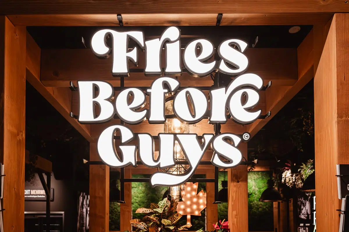 Ein großes, beleuchtetes Schild im Frittenwerk Bielefeld zeigt in fetten, weißen Buchstaben die Worte "Fries Before Guys". Es hängt an einer Holzkonstruktion und wird von sanfter Beleuchtung, Pflanzen und einer grünen Wand umrahmt, die für eine gemütliche Innenatmosphäre sorgen.