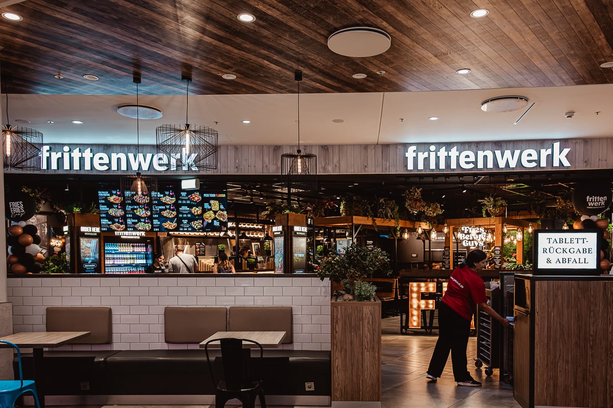 Ein modernes Fast-Casual-Restaurant, Frittenwerk Bielefeld, ist mit Leuchtreklamen ausgestattet. Ein Angestellter in einem roten Hemd hantiert mit Tabletts an der Theke. Digitale Speisekarten, Topfpflanzen, ein Getränkekühlschrank und Pendelleuchten ergänzen die leeren Tische davor, die zum Food Court gehören.