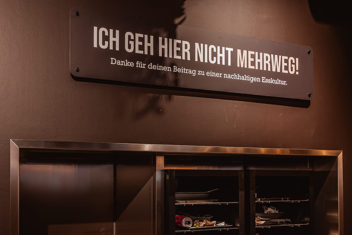 Auf einem Schild an einer braunen Wand im Frittenwerk Bielefeld steht "ICH GEH HIER NICHT MEHRWEG! Danke für deinen Beitrag zu einer nachhaltigen Esskultur." Darunter befinden sich zwei Regale mit Tabletts. Darauf verstreutes benutztes Geschirr und Besteck.