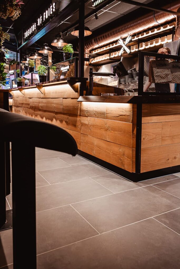 Gezeigt wird eine moderne Restauranttheke, die Abholtheke im Frittenwerk Bielefeld, die aus hellem Holz und schwarzem Metall besteht und über der Hängelampen angebracht sind. Im Hintergrund sind Küchengeräte zu sehen, graue Fliesenböden und ein teilweise sichtbarer Stuhl vervollständigen das saubere, stilvolle Ambiente. Davor ist eine Wartebank zum Anlehnen links zu sehen.