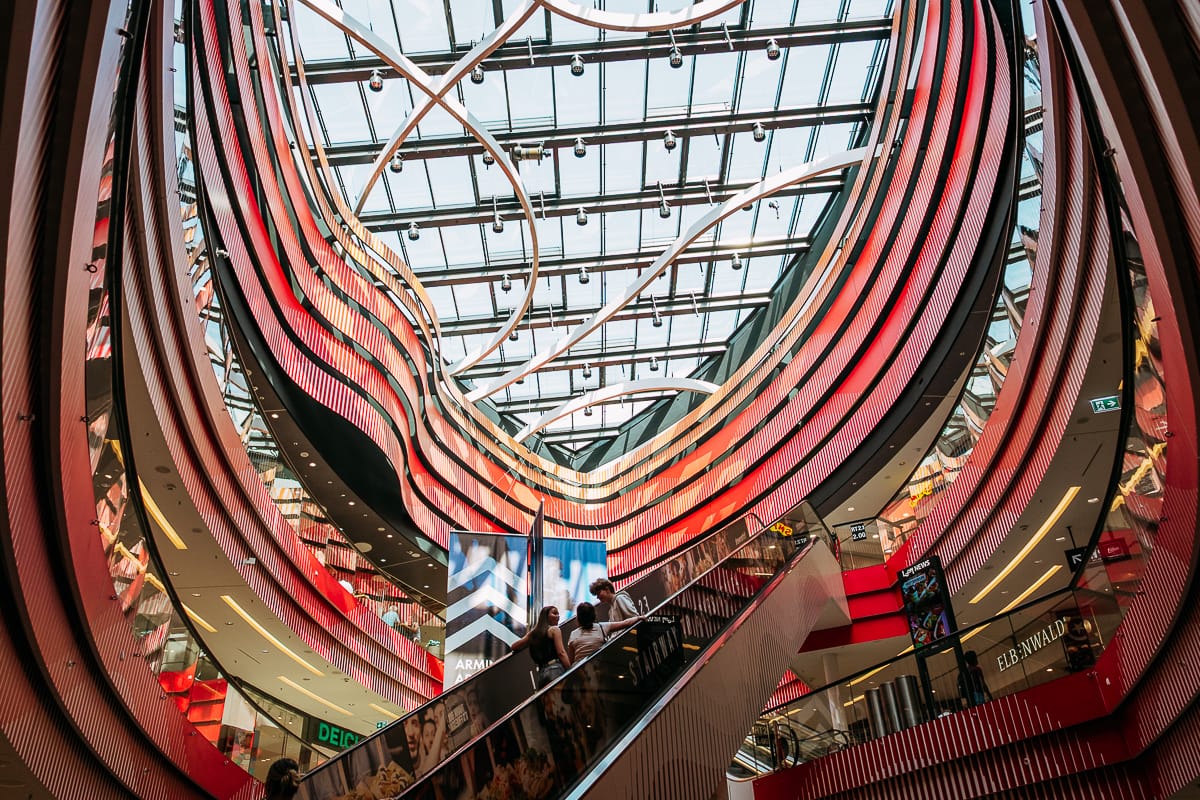 Ein modernes Einkaufszentrum mit geschwungenen roten Wänden, einer Glasdecke und abstrakten Beleuchtungskörpern. Die Menschen fahren auf einer Rolltreppe an Bannern und Ladenschildern vorbei. Natürliches Licht strömt herein und unterstreicht die architektonische Gestaltung.